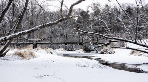 A snowy landscape featuring a wooden bridge over a frozen stream, surrounded by bare trees and winter scenery.