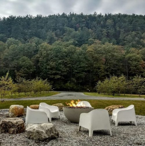 A cozy fire pit surrounded by white chairs, set against a backdrop of trees and a cloudy sky.