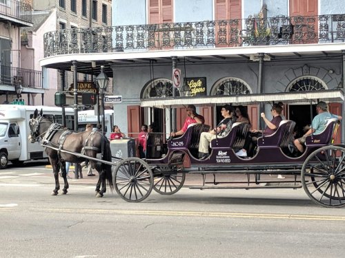 A horse-drawn carriage with passengers rides through a historic street, surrounded by buildings and shops.