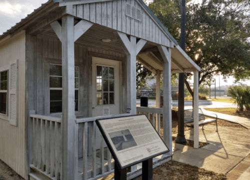 A small wooden building with a porch and a historical marker in front, set in a park with trees in the background.
