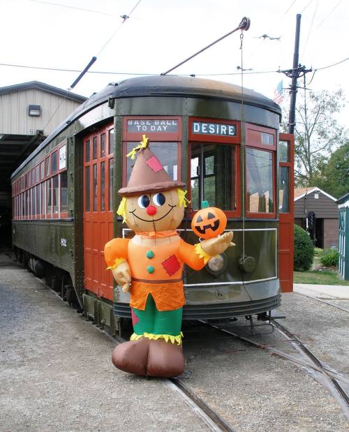 A colorful scarecrow holding a pumpkin stands next to a vintage streetcar with "Desire" on the front.