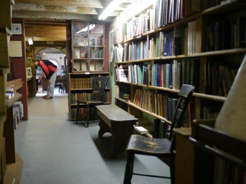 A cozy library aisle lined with bookshelves, featuring wooden chairs and a person browsing in the background.