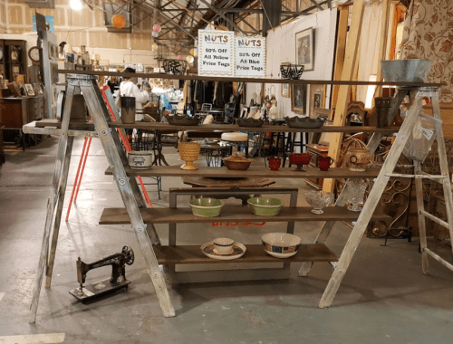 A wooden ladder shelf displays various vintage items, including bowls, vases, and a sewing machine, in a market setting.