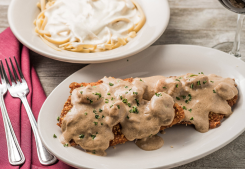 A plate of fried chicken covered in creamy gravy, served with a side of fettuccine alfredo and a glass of red wine.