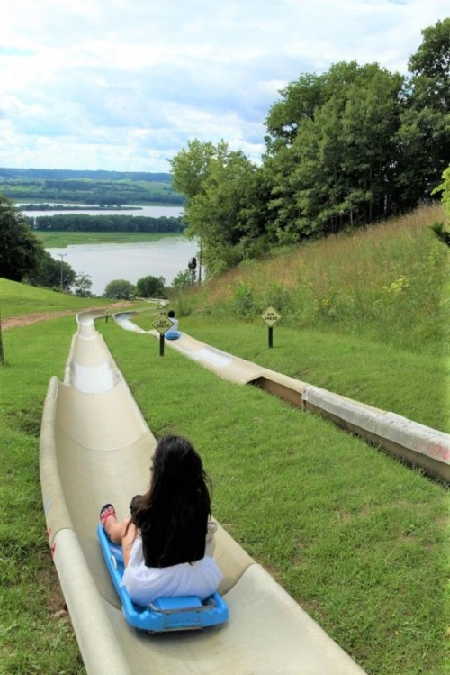 A child sits on a sled, ready to slide down a grassy hill with a scenic view of a river and trees in the background.