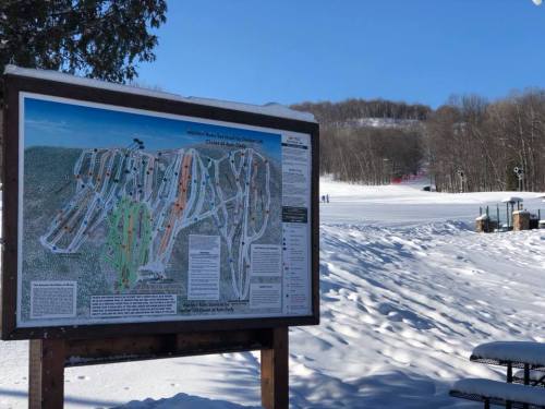 A snowy landscape featuring a ski trail map sign, with slopes and trees in the background.