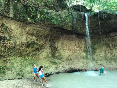 Two people sit by a small waterfall in a forested area, while another person wades in the water nearby.