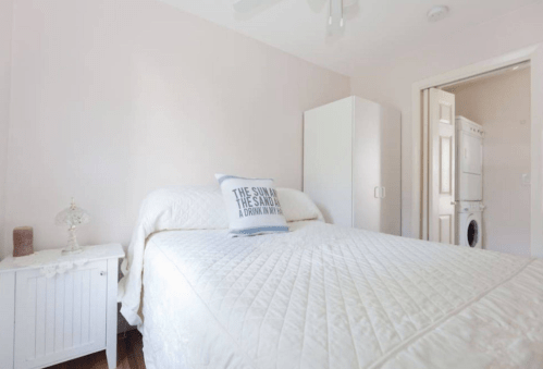 Bright, minimalist bedroom with a white bed, decorative pillow, and a laundry area visible in the background.