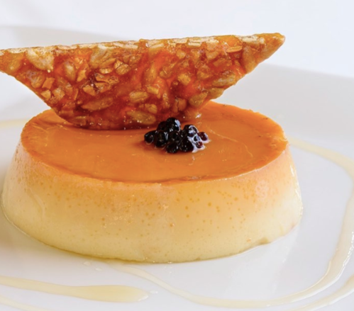 A creamy flan topped with a caramelized crisp and dark berries, drizzled with a light syrup on a white plate.