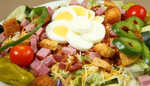 A colorful salad with lettuce, diced ham, cheese, cherry tomatoes, green peppers, croutons, and sliced hard-boiled eggs.