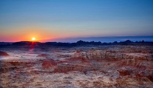 A stunning sunset over a rugged, barren landscape with layered rock formations and distant hills.