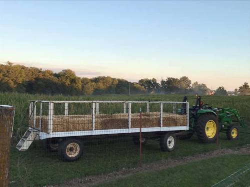 A green tractor pulls a hay wagon in a field, with trees and a clear sky in the background.