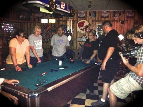 A group of people gathered around a pool table in a dimly lit bar, with one person filming the scene.