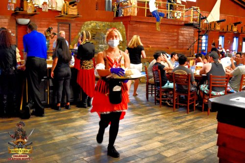 A waitress in a pirate-themed restaurant carries a tray while guests dine at wooden tables in a lively atmosphere.