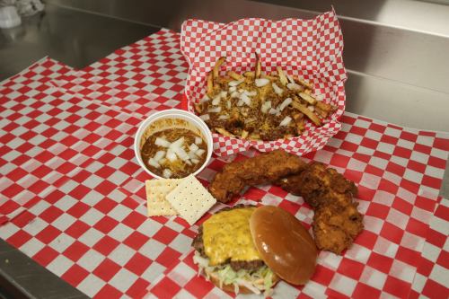 A plate featuring a burger with cheese, fried chicken, fries topped with onions, and a cup of chili with crackers.