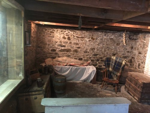 A rustic basement with stone walls, wooden barrels, a rocking chair, and a covered bed. Soft lighting enhances the atmosphere.
