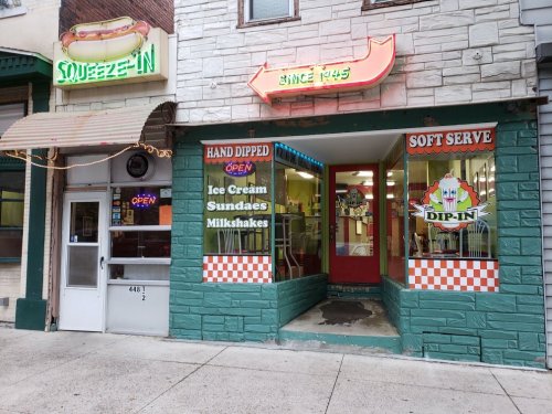 A colorful ice cream shop with a neon sign, featuring a menu of sundaes and milkshakes, located on a city street.