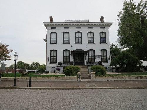 A large, historic white building with black windows and a staircase, surrounded by trees and a cloudy sky.