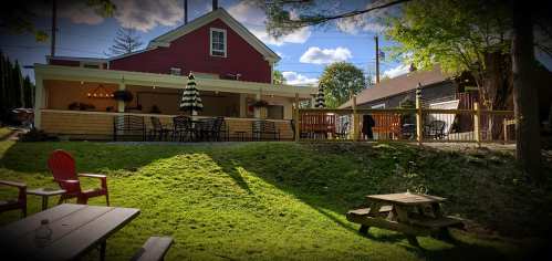 A cozy outdoor seating area with tables and chairs, surrounded by greenery and a red building in the background.