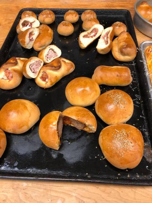 A tray of freshly baked pastries, including stuffed rolls and buns, some with sesame seeds, on a wooden surface.