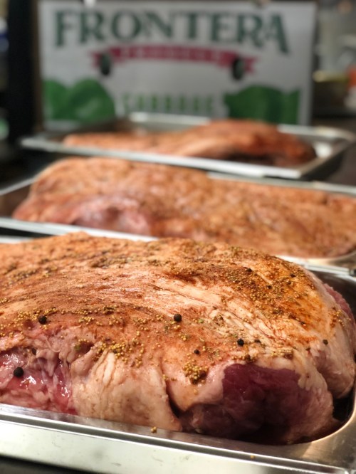 Raw seasoned meat in metal trays, with a blurred background featuring a "Frontera" sign.