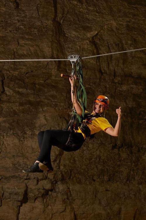 A person in a yellow shirt and helmet zip-lining in a cave, giving a thumbs-up with a smile.