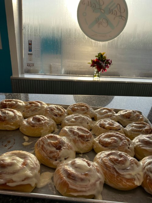 A tray of freshly baked cinnamon rolls with cream cheese frosting, set against a foggy window and a small flower arrangement.
