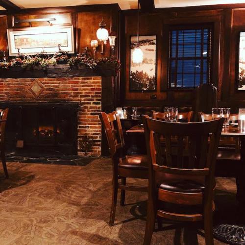 Cozy restaurant interior with wooden tables, chairs, a brick fireplace, and festive decorations on the walls.