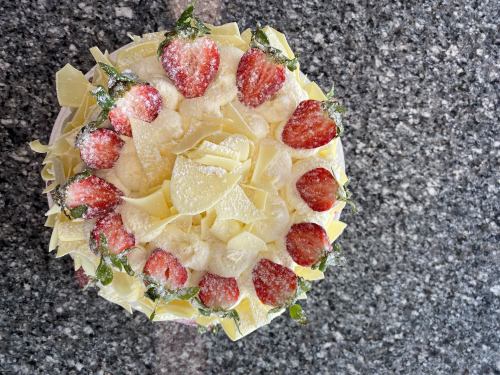 A round cake topped with strawberries, white chocolate shavings, and whipped cream, set on a granite surface.