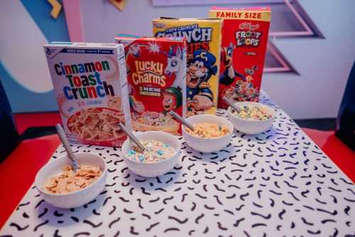 A table with four bowls of cereal and colorful cereal boxes, including Cinnamon Toast Crunch and Lucky Charms.