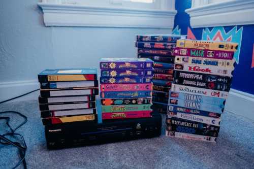 A collection of VHS tapes stacked beside a VCR, with titles like "Teenage Mutant Ninja Turtles" and "Jurassic Park."