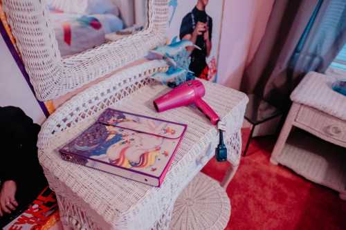 A wicker vanity table with a pink hairdryer and a colorful unicorn-themed binder on top, set in a cozy room.