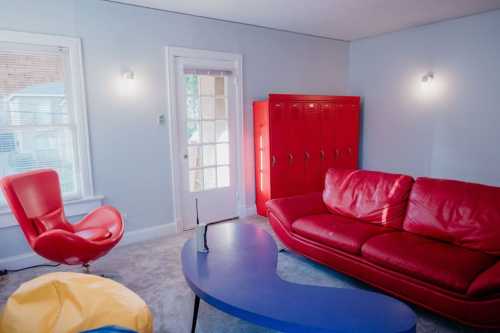 A bright room featuring a red couch, colorful chair, blue table, and red lockers, with natural light from a window.