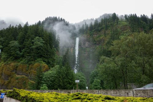 A tall waterfall cascades down a lush, green hillside surrounded by trees and misty clouds.