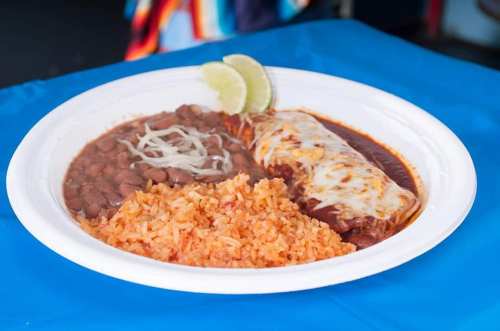 A white plate with enchiladas topped with cheese, rice, refried beans, and lime wedges on a blue table.