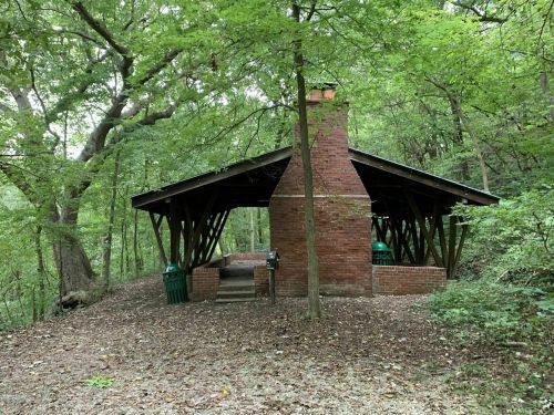 A rustic brick shelter with a chimney, surrounded by lush green trees and a gravel path.