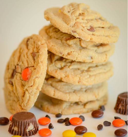 A stack of peanut butter cookies with chocolate chips, surrounded by Reese's Pieces and mini peanut butter cups.