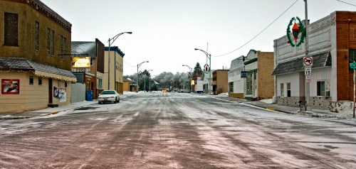 Snow-covered street in a small town, lined with buildings decorated for the holidays, under a gray sky.