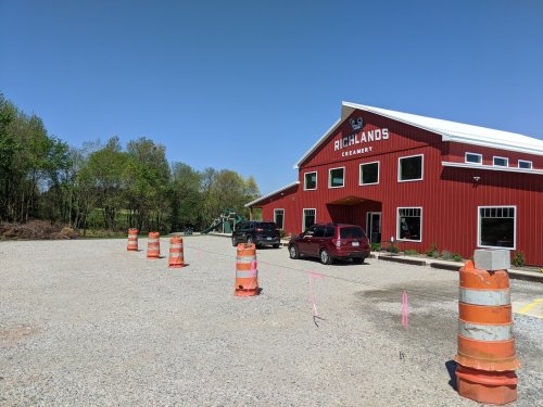 A red barn-style creamery with parked cars and construction cones in a gravel lot, surrounded by trees and blue sky.