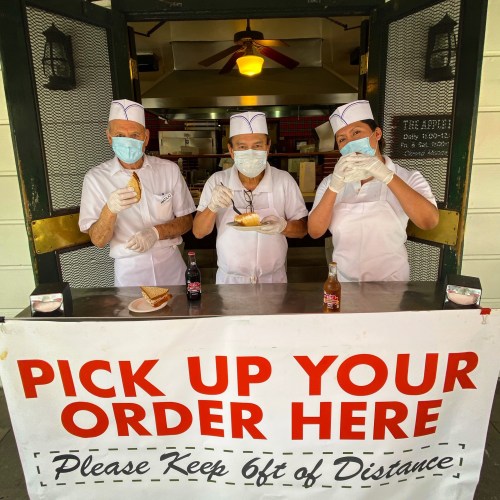 Three masked workers in white uniforms hold food items at a takeout window, with a sign reading "Pick Up Your Order Here."