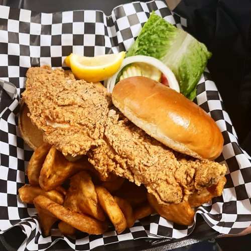 A plate with a large fried chicken sandwich, lettuce, tomato, lemon wedges, and crispy potato wedges in a checkered basket.