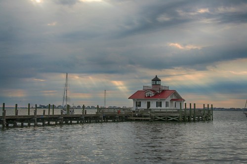 A lighthouse on a pier under a cloudy sky, with rays of light breaking through the clouds over the water.