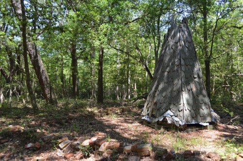 An abandoned, conical wooden structure surrounded by trees and overgrown vegetation in a forested area.