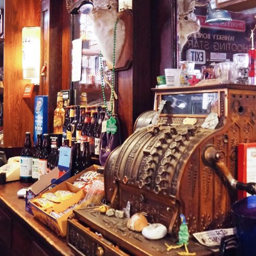 A vintage cash register sits on a bar counter, surrounded by bottles and snacks, with a deer head mounted on the wall.