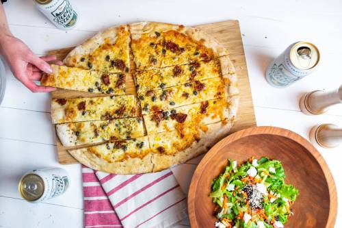 A hand reaches for a slice of pizza on a wooden board, with a salad and drinks nearby.