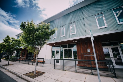 Modern building exterior with large windows, wooden accents, and trees lining the sidewalk under a partly cloudy sky.