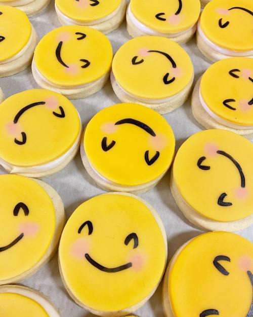 A close-up of yellow smiley face cookies with pink blush and black icing details, arranged on a white surface.
