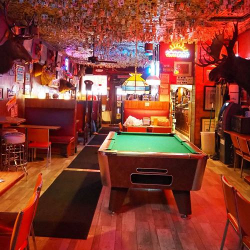 A vibrant bar interior with a pool table, colorful neon lights, and walls adorned with dollar bills and mounted animal heads.