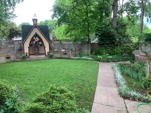 A serene garden with a small chapel, lush green grass, and a winding stone path surrounded by trees and plants.