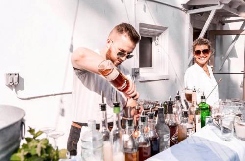 A bartender pours a drink at an outdoor bar while a smiling woman watches, surrounded by various bottles.